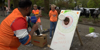 Rudo gooit in de Rudo! XL Koningsdag aflevering een bord slagroom naar een jongen die zijn hoofd door een bord steekt. Dit is een van de activiteiten op de vrijmarkt in Wassenaar.