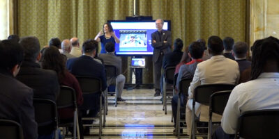 De twee initiatiefnemers van het traject staan voor een scherm tijdens een presentatie. Voor hen zit de grote groep statushouders.