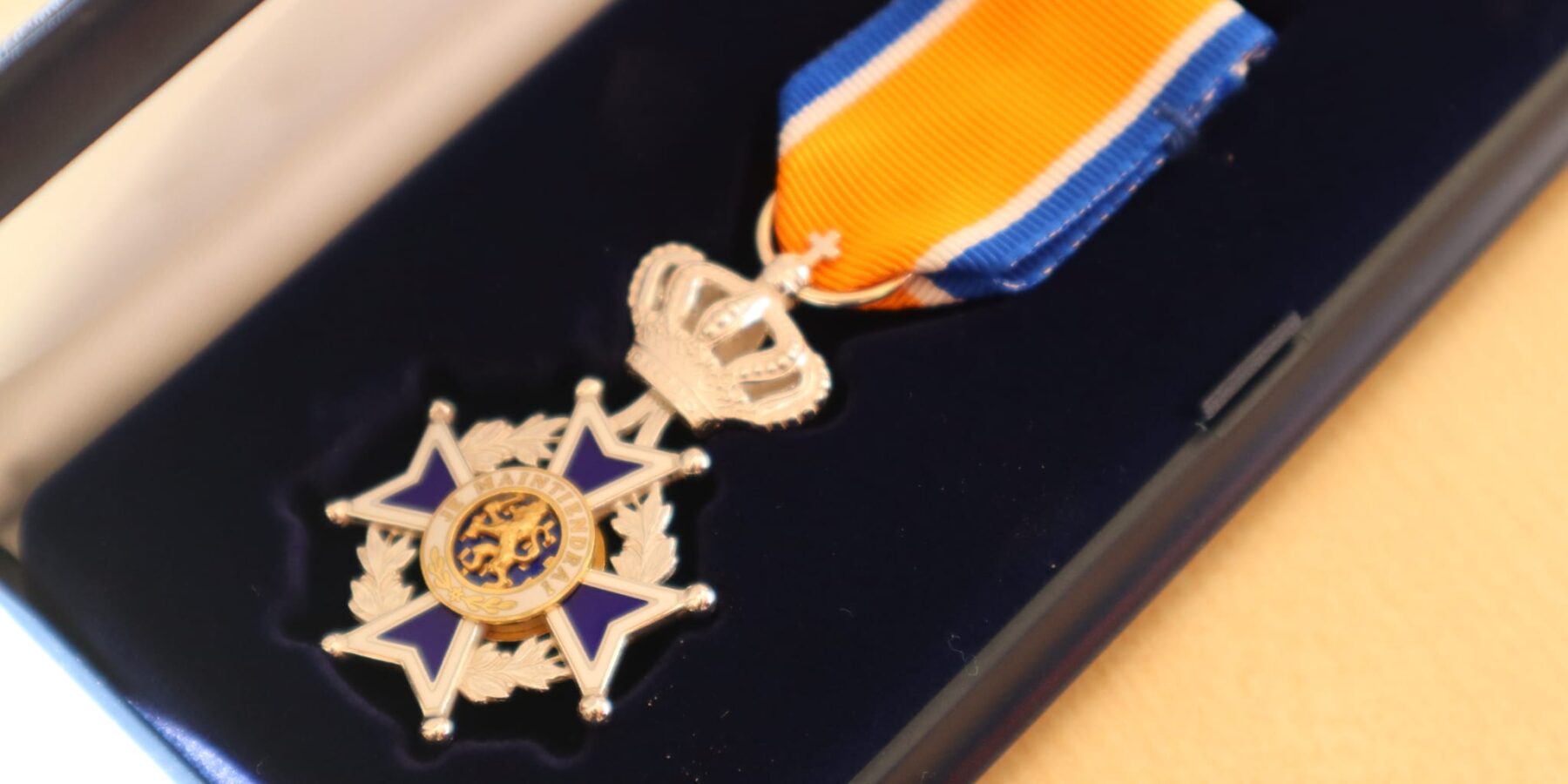 Op de afbeelding is een Koninklijke Onderscheiding te zien. Het is een blauw-oranje lintje met een broche in de vorm van een kroon.