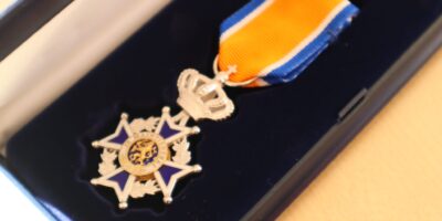 Op de afbeelding is een Koninklijke Onderscheiding te zien. Het is een blauw-oranje lintje met een broche in de vorm van een kroon.