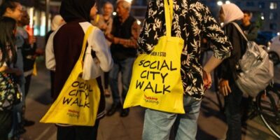 Deelnemers aan de Social City Walk staan met gele draagtassen in gesprek tijdens een bijeenkomst in de avonduren.