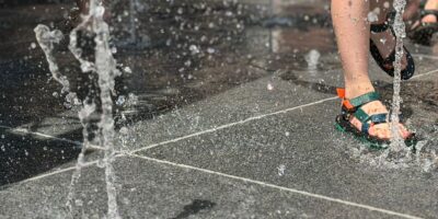 Water dat uit de waterfontein komt met voeten van kinderen die aan het spelen zijn. De Waterfontein in Wassenaar is buiten gebruik door hitte. Reparaties worden uitgesteld tot betere weersomstandigheden.
