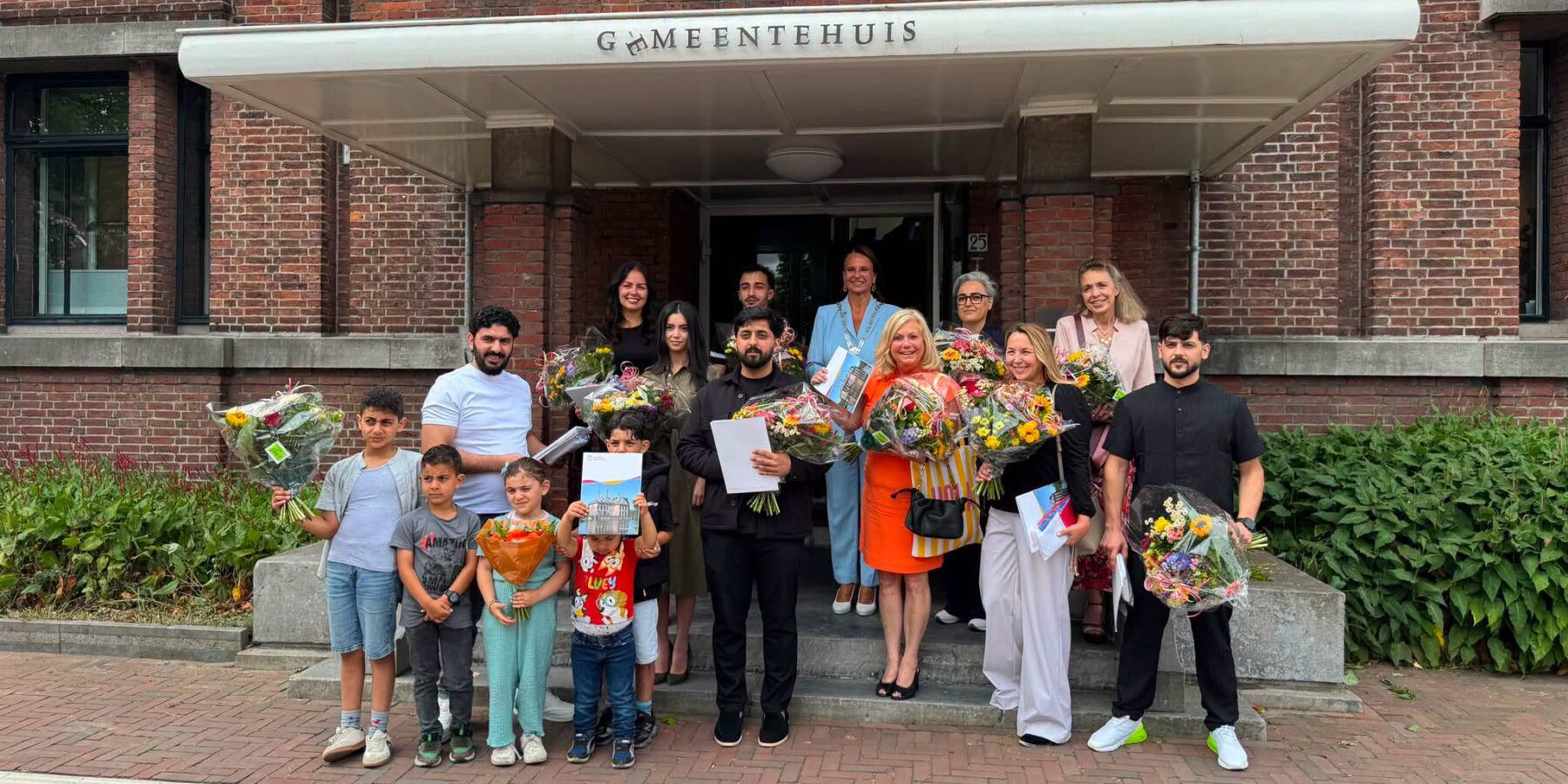Voorschoten viert naturalisatie 15 nieuwe Nederlanders. Ze poseren met burgemeester Nadine Stemerdink met bloemen op de trap voor het gemeentehuis.