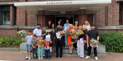 Voorschoten viert naturalisatie 15 nieuwe Nederlanders. Ze poseren met burgemeester Nadine Stemerdink met bloemen op de trap voor het gemeentehuis.